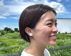 Woman with short hair wearing a white earring outdoors with greenery and blue sky in the background