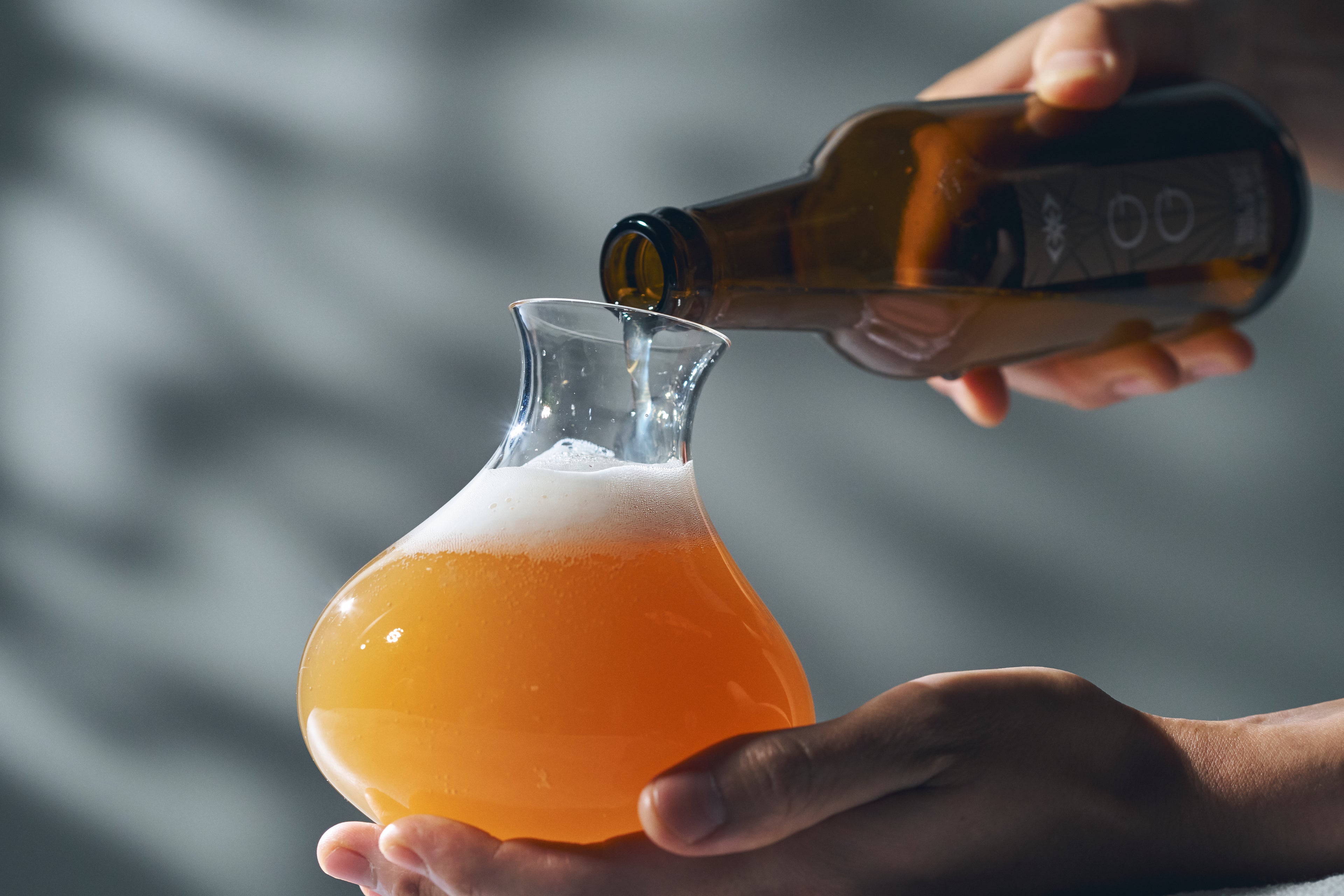 Action shot of golden craft beer being poured from a bottle into a bulbous hand-blown glass, creating a fresh foam head.