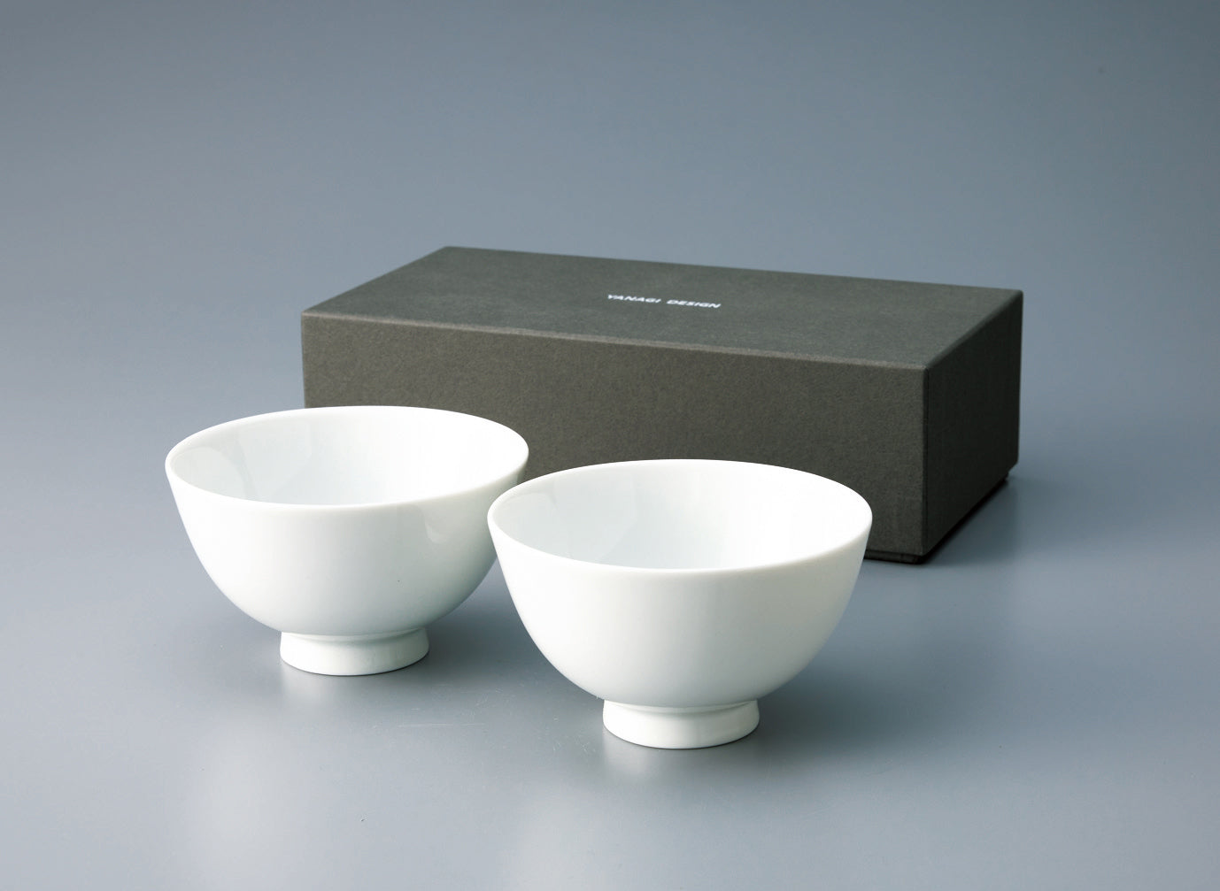 Pair of **plain white Japanese porcelain rice bowls** with a footed base, displayed in front of the dark gray retail gift box.
